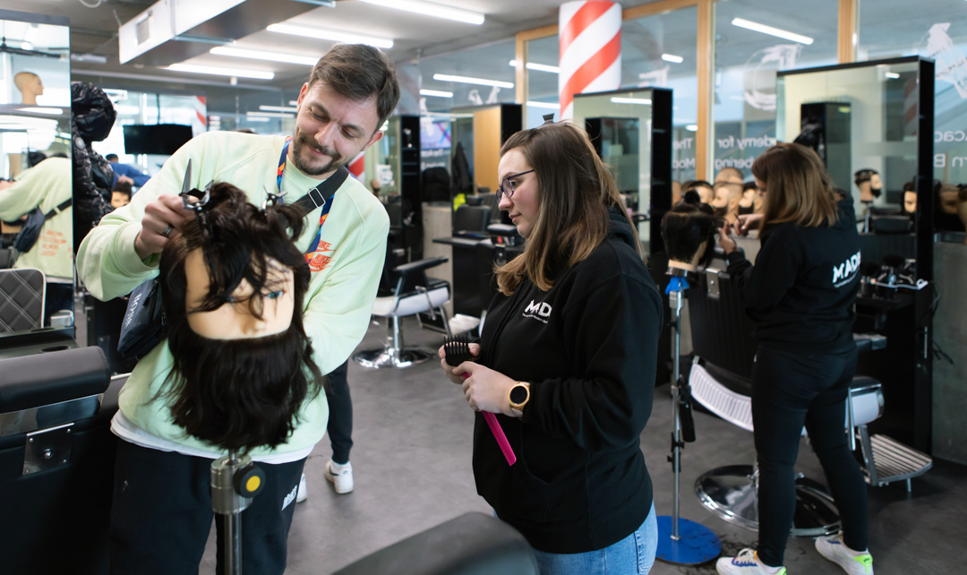 A male instructor in a light green sweatshirt assisting a female student with a mannequin head in a barbering classroom.