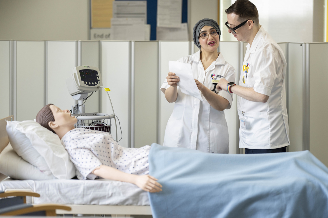 Two care students discuss notes at a hospital bed during a training exercise