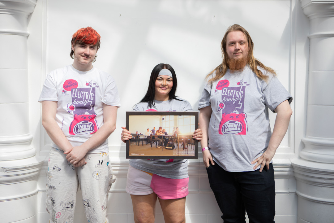 Three students wearing Electric Honey t-shirts standing in front of a white wall, holding a framed classroom photo from 2008.