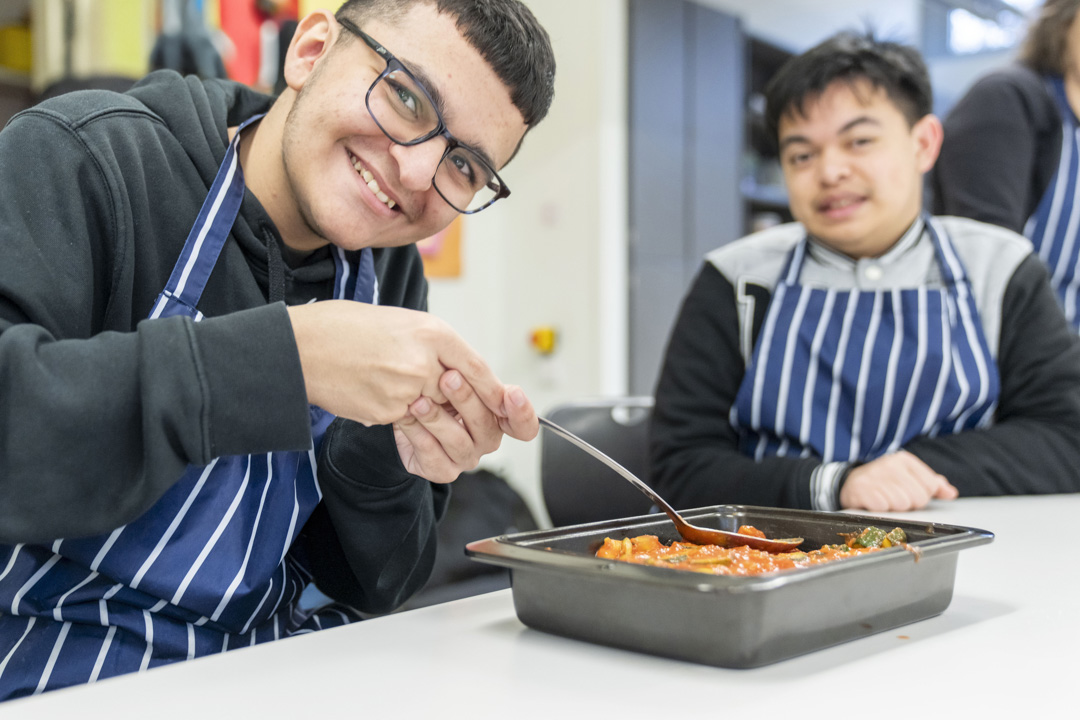 Two young men smiling and preparing a tray of food, wearing striped aprons in a kitchen setting.