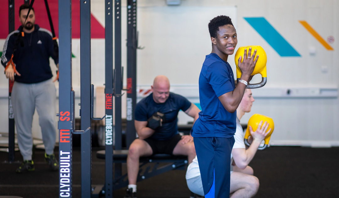 Student smiling while holding a yellow kettlebell during a strength training session at the gym.