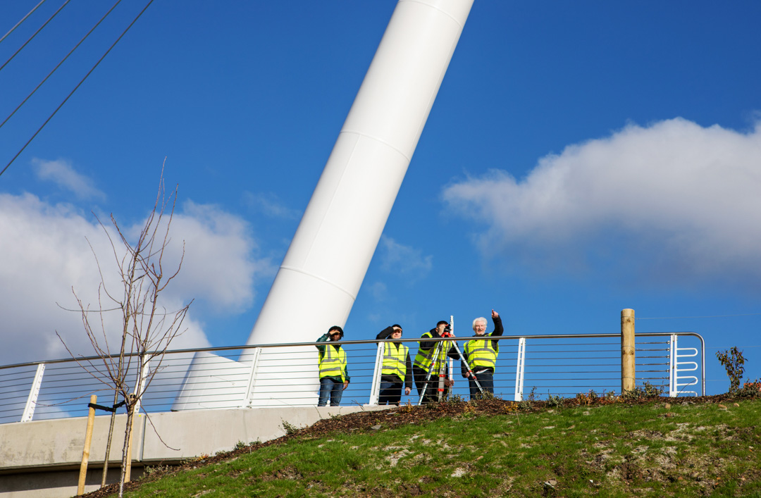 Wide-angle view of the Stockingfield Bridge with a group of students and lecturers in high-visibility vests conducting a surveying exercise on the platform beneath a bright blue sky.