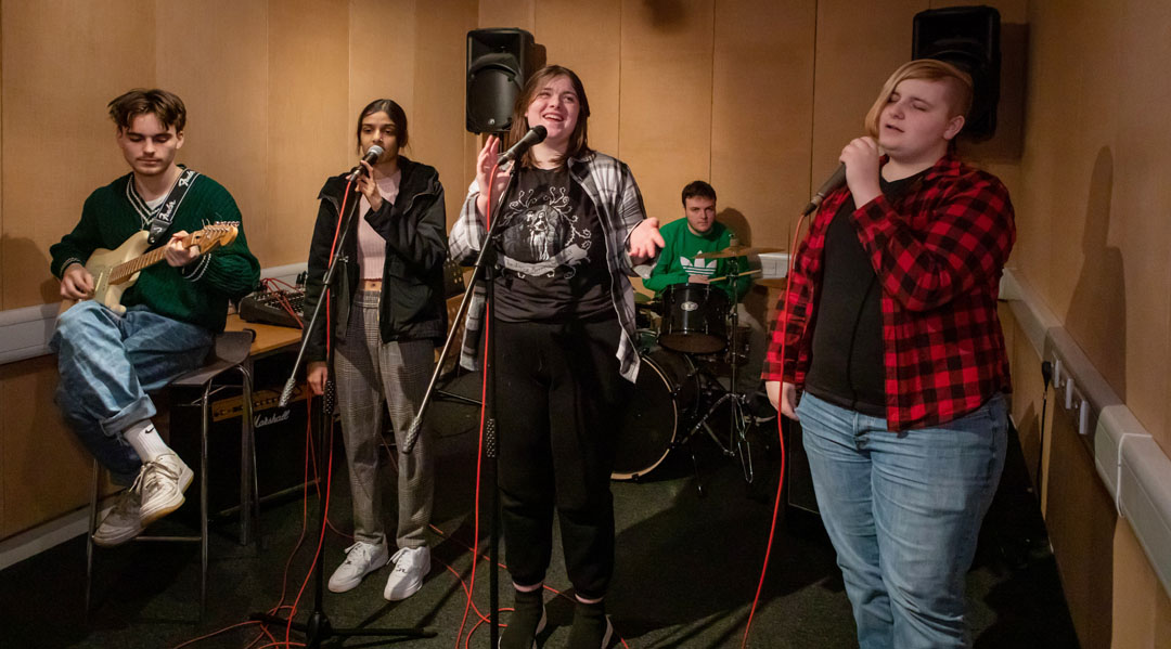 A group of five students rehearsing, featuring a guitarist, two vocalists, a bassist, and a drummer in a music studio.