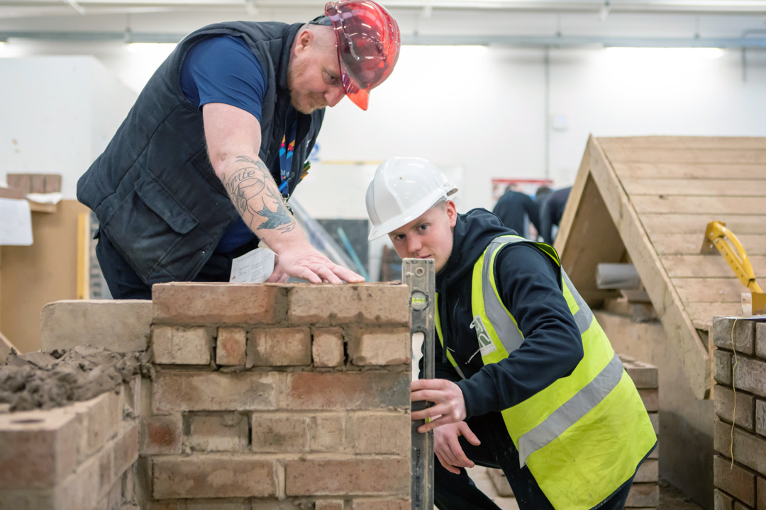 A brickwork lecturer guiding a student as they carefully check alignment and leveling of a brick wall construction in a practical workshop.
