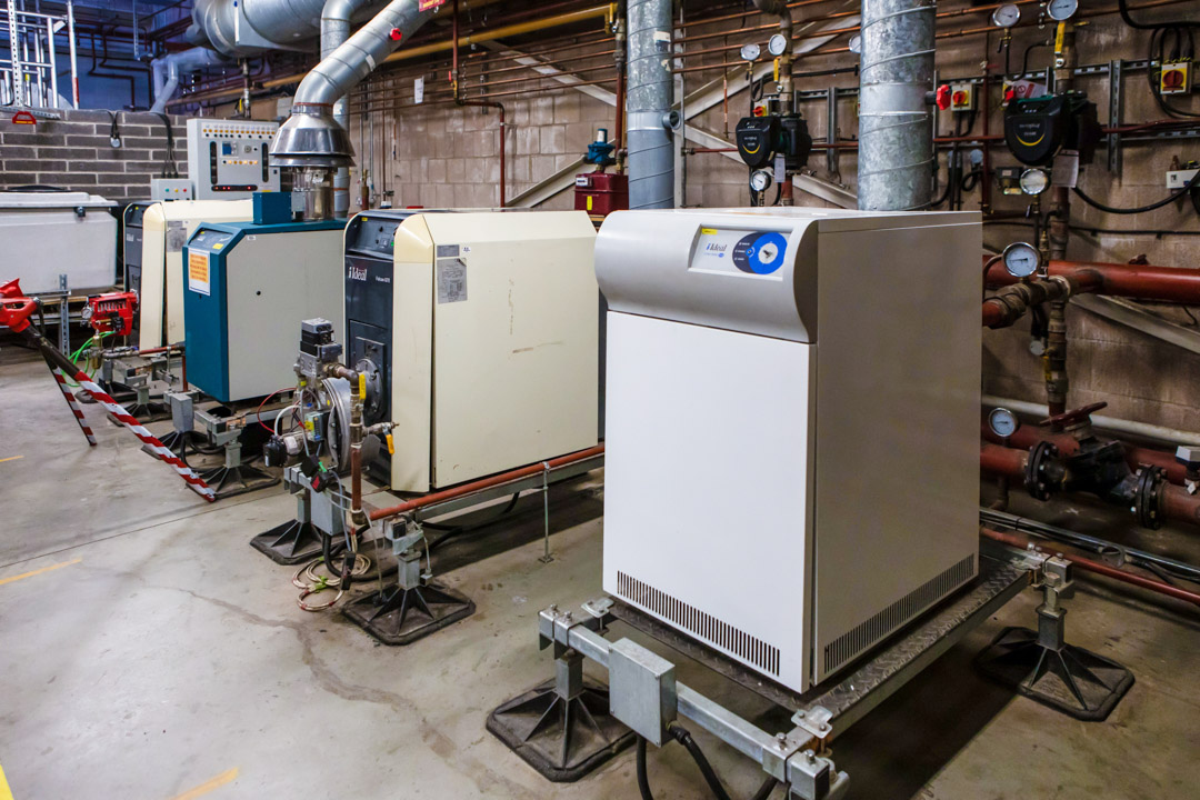 A row of industrial boilers in a building services training facility, connected to ventilation ducts and a complex piping system.
