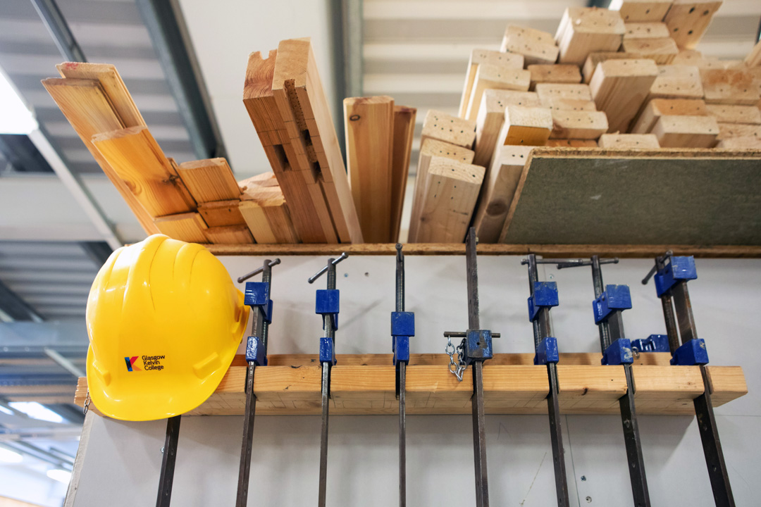 Woodworking equipment including clamps, timber planks, and a bright yellow safety helmet neatly arranged on a storage rack in a joinery workshop.