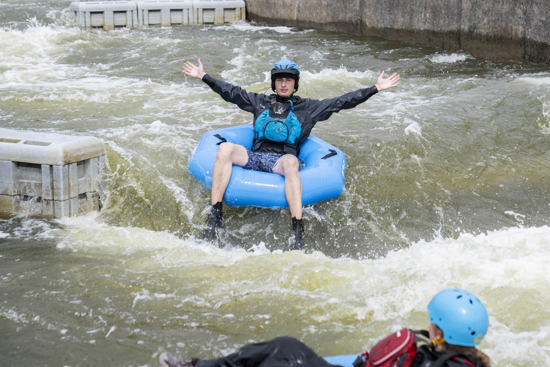 A student with arms outstretched riding a blue inflatable ring on a whitewater course.
