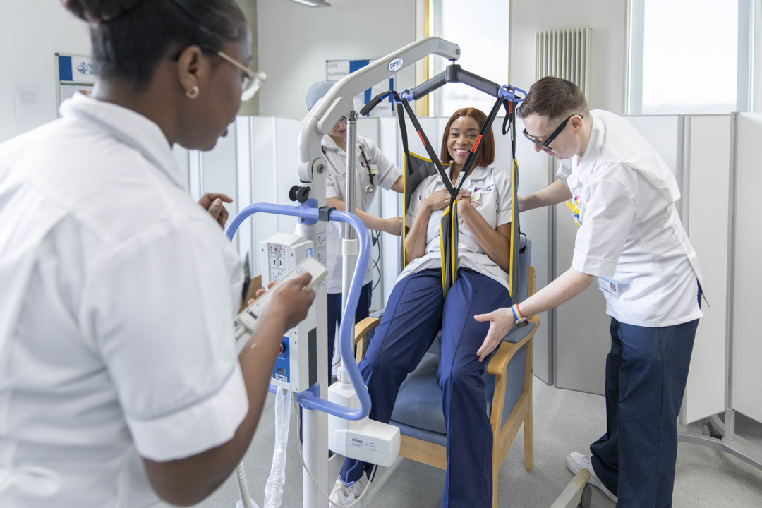 Care students practice using a patient hoist with a seated classmate in a simulated care setting.