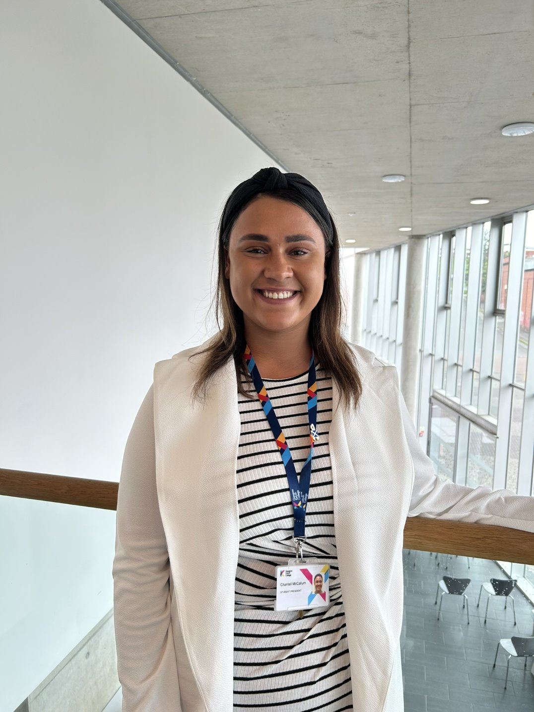 A woman in a white blazer and striped dress stands near a railing in a modern building with large windows. She smiles warmly at the camera, wearing a lanyard with an ID badge.