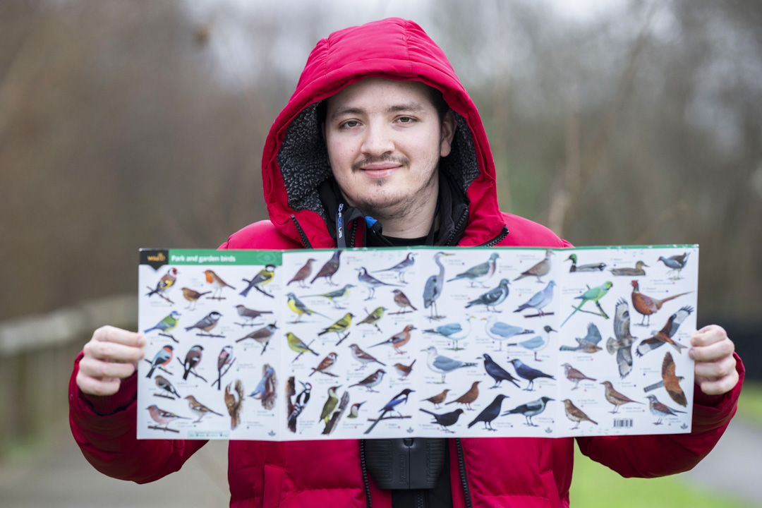 Smiling man in a red jacket holding a bird identification poster with various bird species displayed.