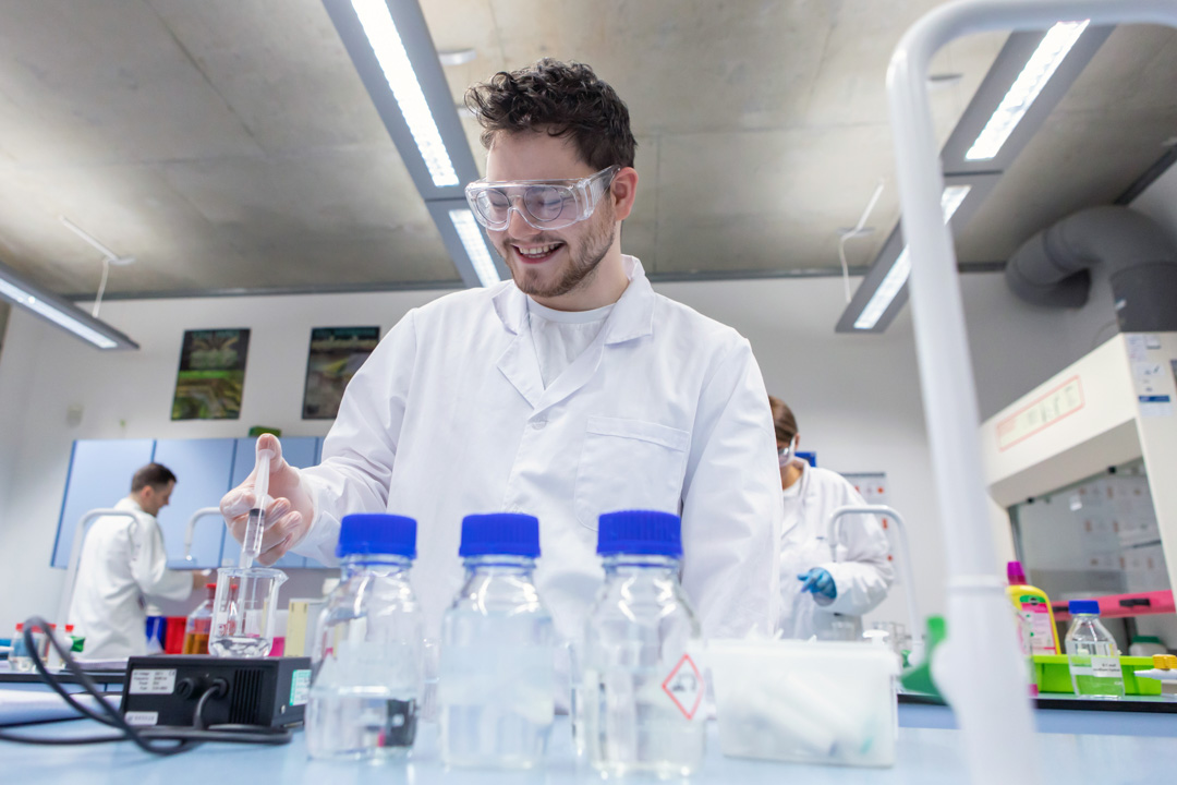 Male student wearing lab coat and goggles pipetting liquid during science experiment