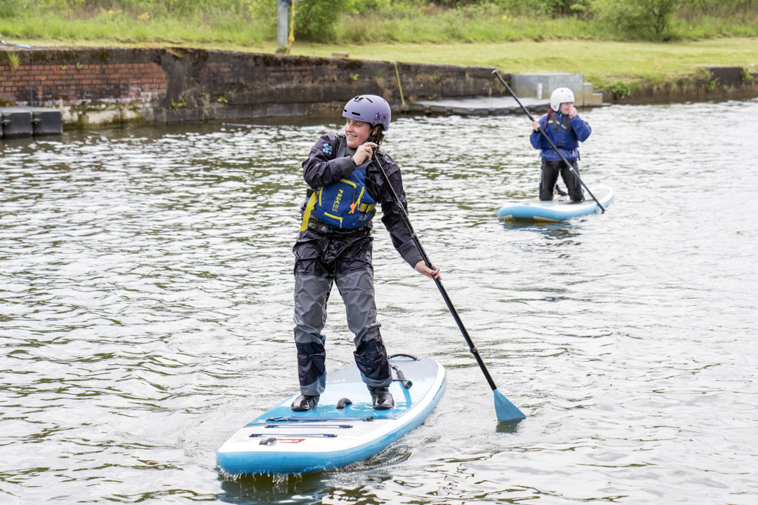 Two people stand-up paddleboarding on a calm canal, both wearing helmets and buoyancy aids.
