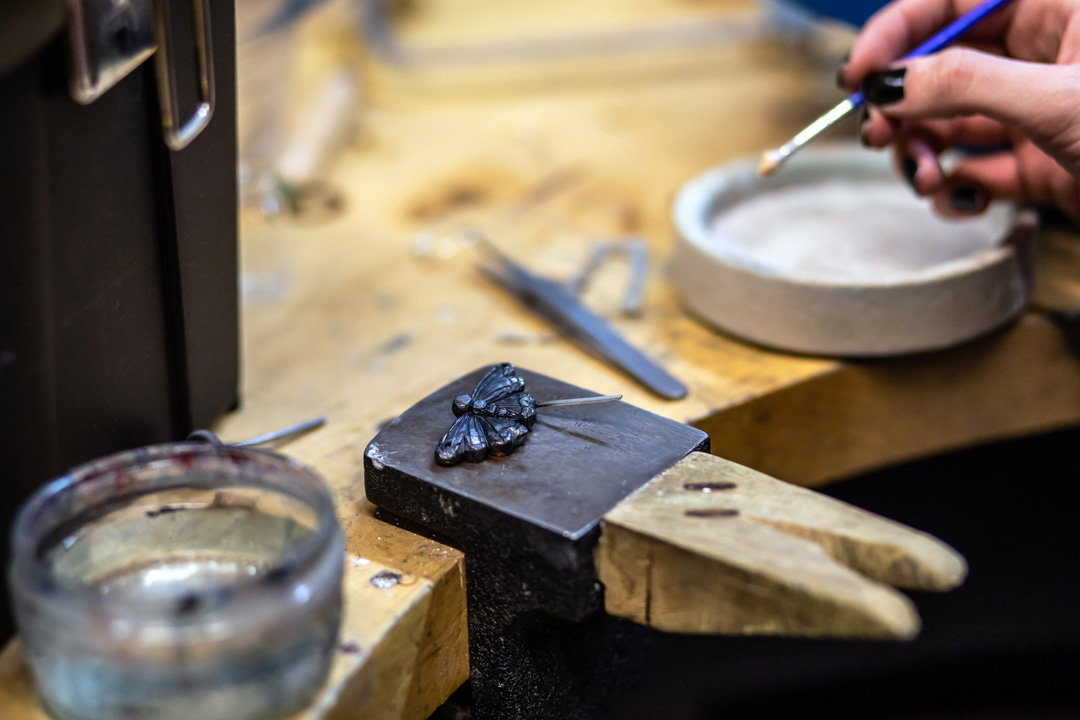 A close-up of a dark metal butterfly-shaped jewellery piece on a metal block, with an artist’s hand holding a fine brush to apply detail work.