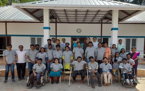 Group photo of staff and youth with disabilities outside a building at IOCL Kerala, with some individuals seated in wheelchairs and others standing, all smiling.