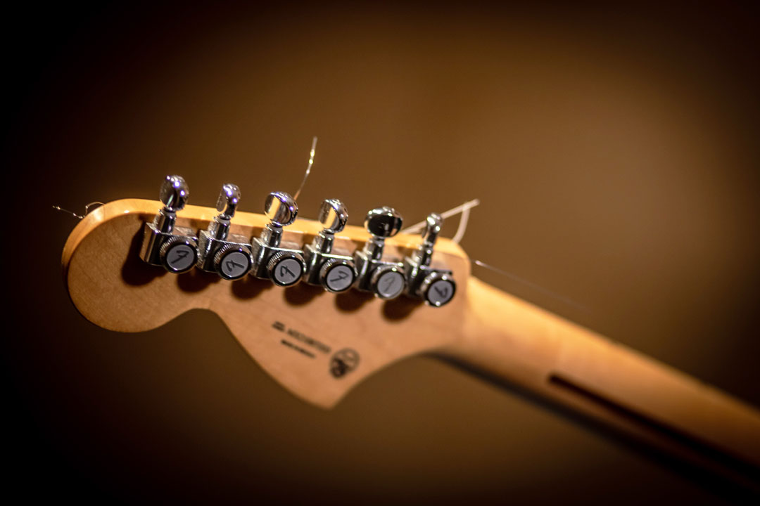 A wooden guitar headstock with chrome tuning pegs and loose strings, set against a blurred background.
