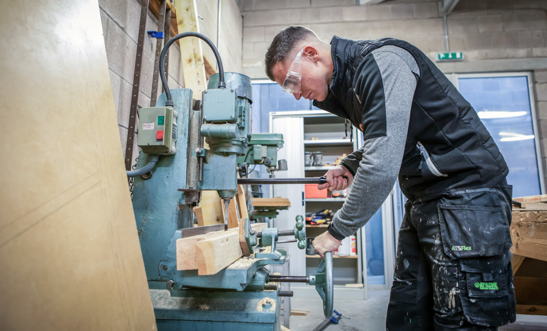 A joinery student wearing safety glasses, operating woodworking machinery to shape timber in a college workshop.