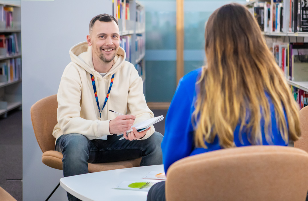Glasgow Kelvin College staff member in a cream hoodie speaking with a student in a library setting, both seated and engaged in conversation with a notepad.