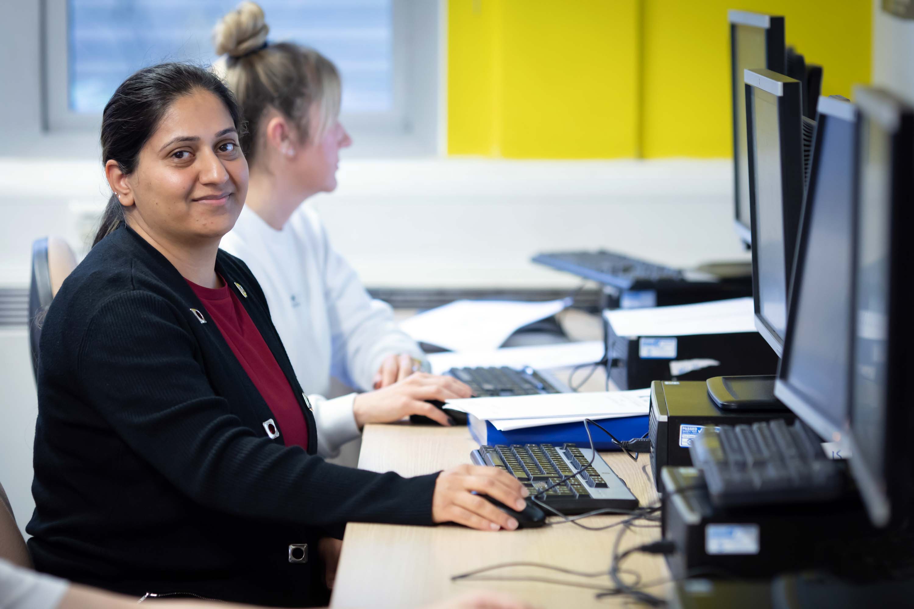 Student in a black cardigan sitting at a computer workstation, smiling while working on a task in a business and admin class.
