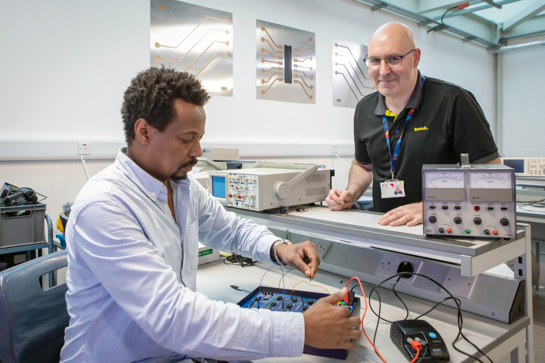 A lecturer and a student in an electronics lab working on a circuit board experiment. The lecturer is smiling while overseeing the student’s work.