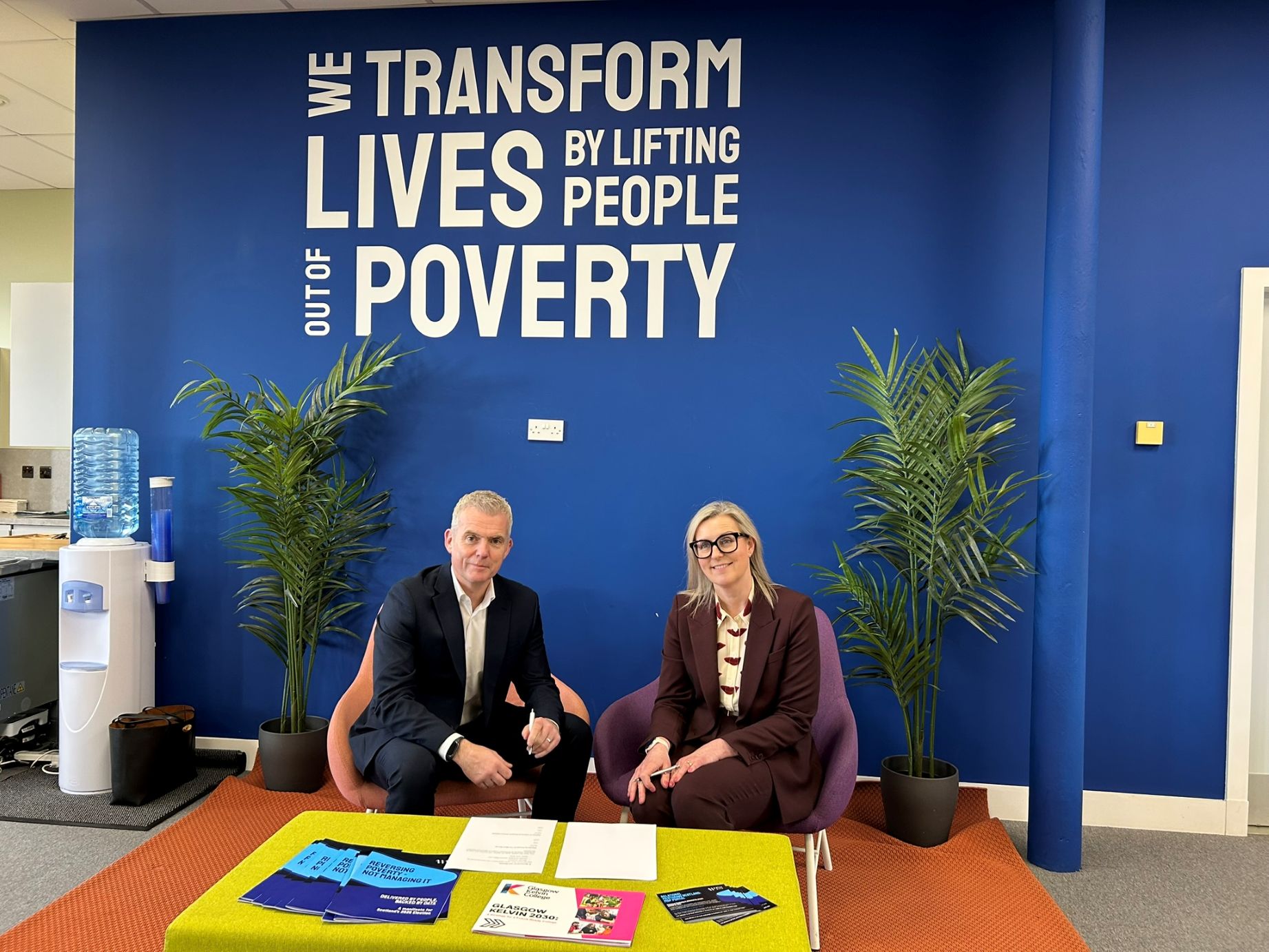 Two professionals seated beneath a “We Transform Lives by Lifting People Out of Poverty” wall, marking a partnership agreement.
