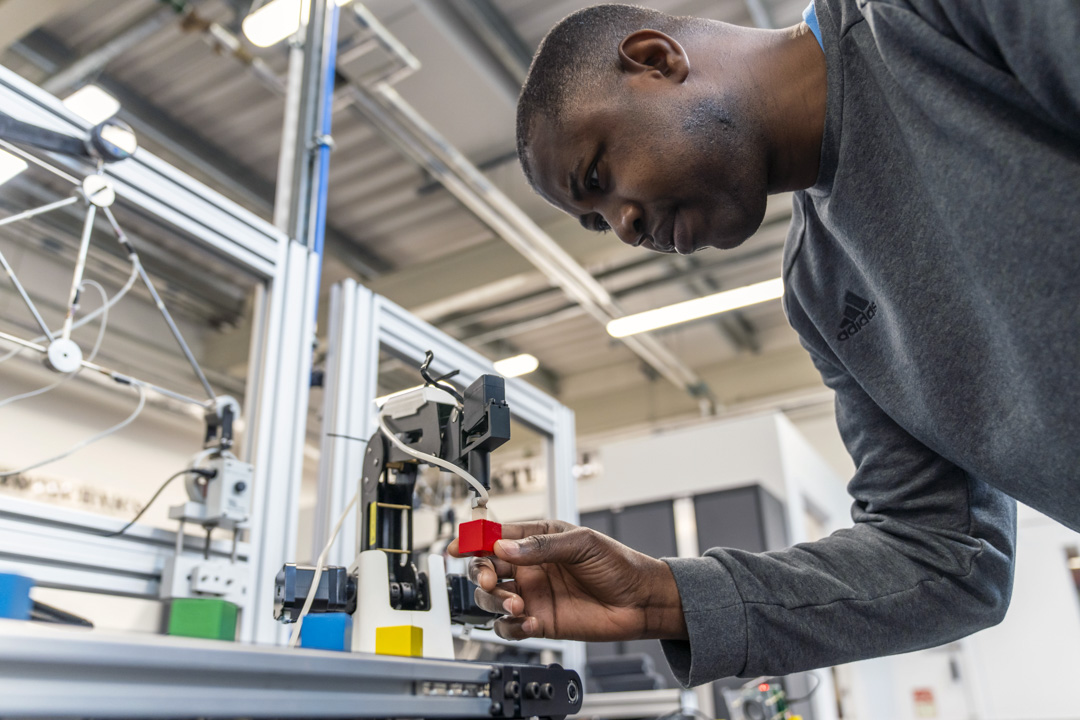 Student adjusting a robotic arm to pick up a red block, focusing intently on precision in a mechatronics workshop.