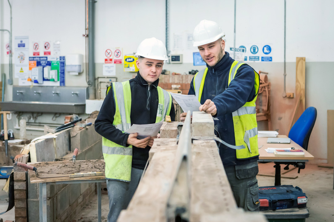 Two brickwork students in safety gear reviewing plans together, standing beside a brick wall and discussing their next steps in a practical training workshop.