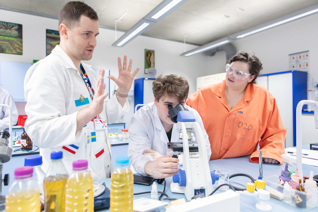 Science lecturer guiding a student on microscope use with classmates observing in the lab at Glasgow Kelvin College