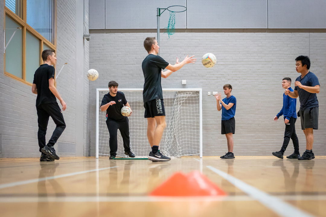 Students in a games hall tossing netballs in a circle as part of a group activity.