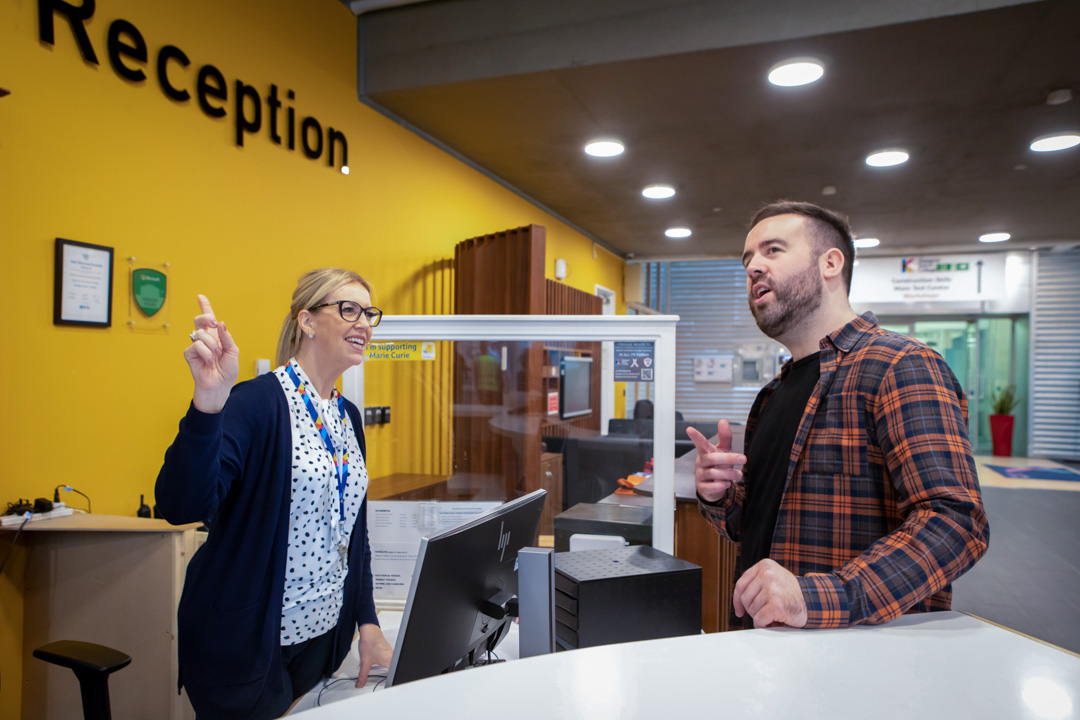 Receptionist assisting a student at the Glasgow Kelvin College Springburn Campus reception desk, both smiling and engaged in conversation.