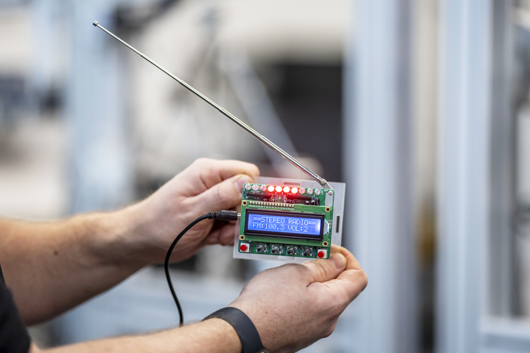 Close-up of a small electronic device with an LCD screen displaying radio frequency settings, held by a person in a technical lab.