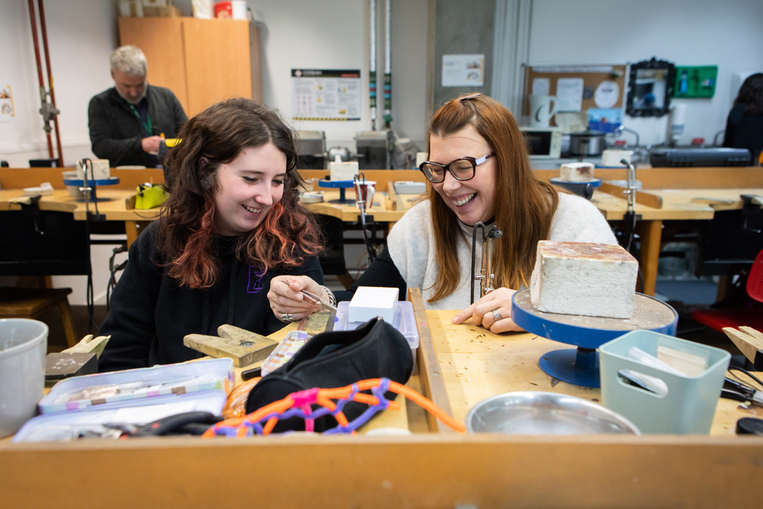 Two women in a jewellery workshop laughing and working together at a wooden workbench, with various tools and equipment scattered around.