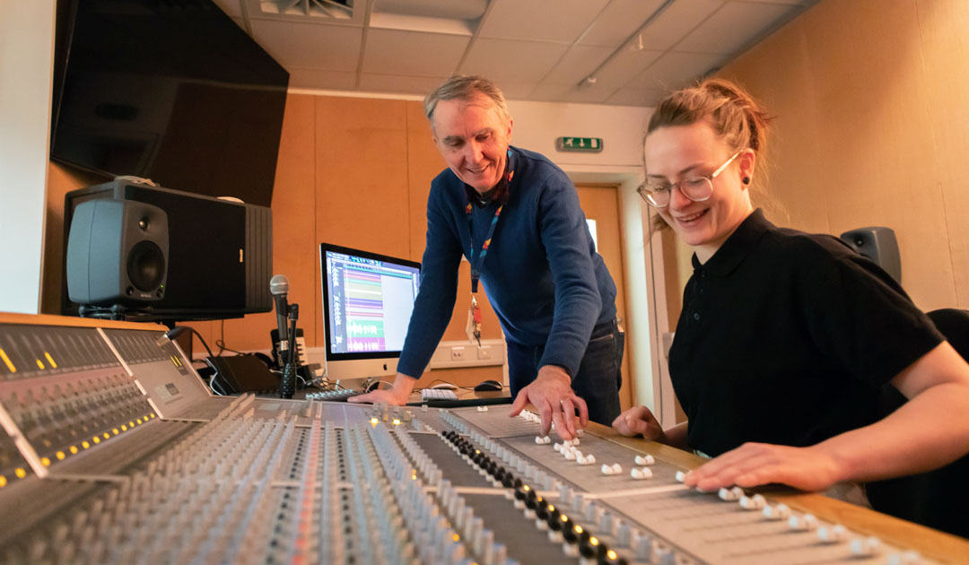 A lecturer and student working together at a large mixing console, discussing adjustments to the sound levels.