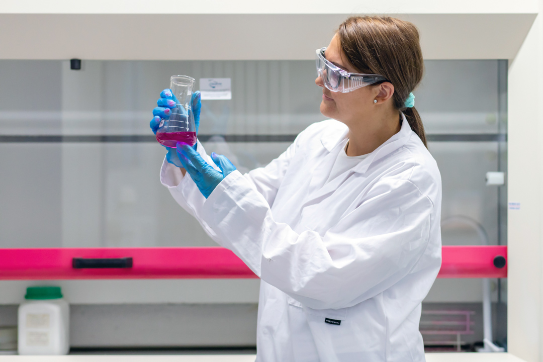Student in a lab coat studying a beaker of pink liquid under bright lighting in a science lab.
