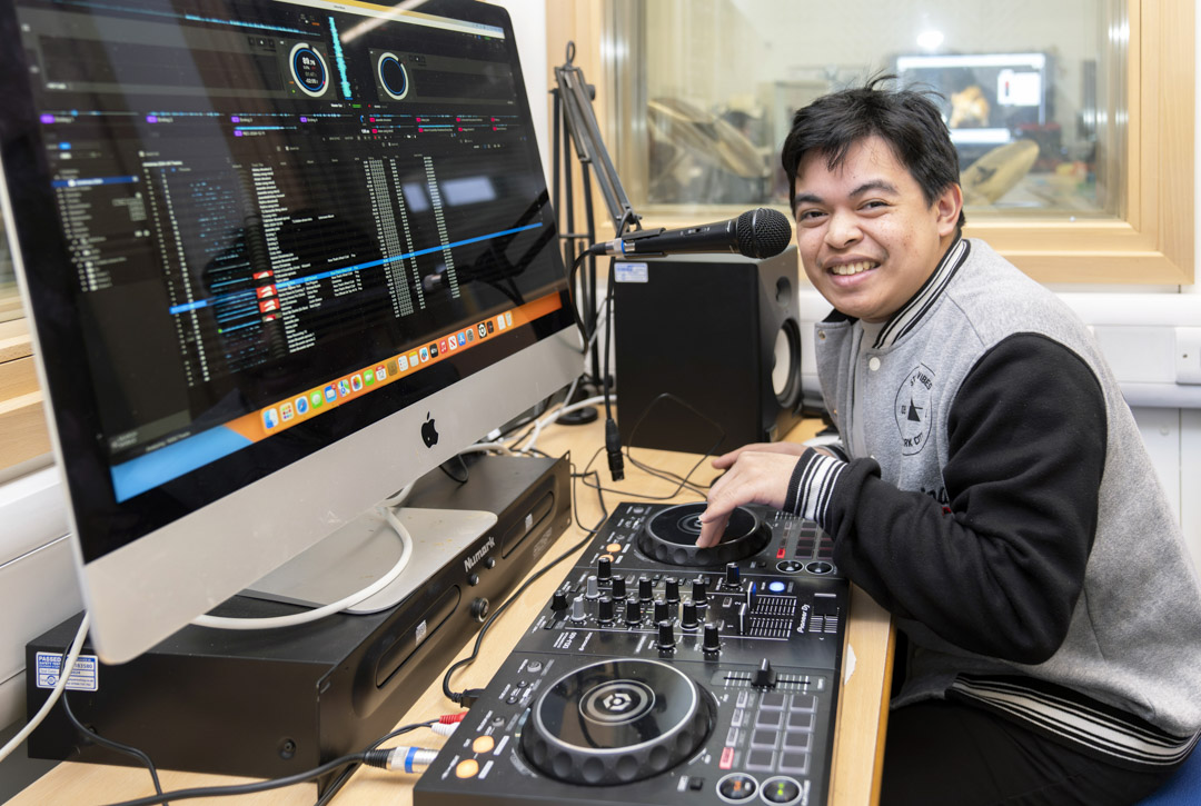 Young man smiling while using DJ equipment and a microphone, with a computer screen displaying music software.